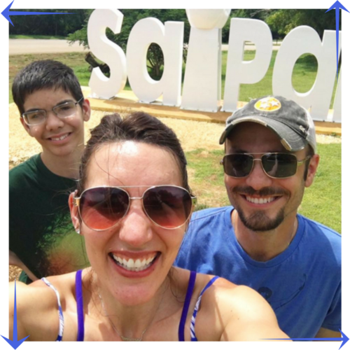 A teenage boy wearing glasses stands with his mother and father in front of a sign that says SAIPAN.